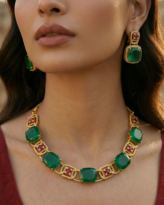 Woman wearing a gold necklace with green and red gemstones, set against a blurred background.
